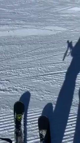 The video shows a person skiing on a snowy slope under a clear blue sky. The camera is attached to the skier, providing a first-person perspective. The beginning of the video shows the person’s skis on the ground, and their shadow as they start to move. The scene transitions to the skier going downhill. Other skiers are visible in the distance on the slope, and a mountainous landscape stretches out behind them. The snow is groomed with visible lines from a snow machine.