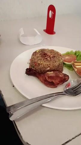 The video shows a table setting at what appears to be a restaurant or cafe. The first shot focuses on a plate of food, consisting of fried rice, a piece of fried chicken, a garnish of lettuce and tomato, and a small dish of chili paste. There is also a fork and spoon on the plate. The second shot pans to show a golden dipping sauce dish and a red drink in a glass. A green toy is also visible on the table. The third shot shows a cup of coffee with latte art, another drink in a glass, and the green toy is still present. The fourth shot shows another plate of food with rice, meat, and crackers, along with a purse in the background. The final shot shows a man standing behind a bar, possibly a barista, in the background of the restaurant. The overall environment appears to be indoors with soft lighting and modern decor.
