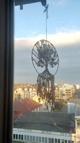 From an elevated, indoor perspective, a macrame tree-of-life decoration hangs suspended against a backdrop of a European cityscape under a partly cloudy sky. The tree-of-life is crafted from interwoven rope and adorned with small, spherical objects that could be beads or stones. Below, various buildings with mixed architectural styles spread across the landscape, including buildings with flat roofs and others with peaked, red-tiled roofs. A crane is visible in the distance. The footage is framed by a window, suggesting the camera is positioned indoors, and the view is slightly obscured by the window frame.