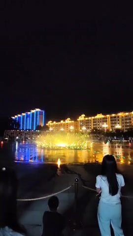 The video shows a night scene with a fountain show in front of illuminated buildings. Three people are visible in the foreground, two women and one child, facing away from the camera towards the fountain. The woman on the left is wearing a white shirt and checkered pants, with a white bag slung over her shoulder. The child, wearing a dark shirt, is crouching. The woman on the right is wearing a white shirt and white pants. The fountain is illuminated with yellow and green lights, with water jets creating a dynamic display. The buildings in the background are lit up in blue and yellow. The ground in front of the fountain appears wet, reflecting the lights.