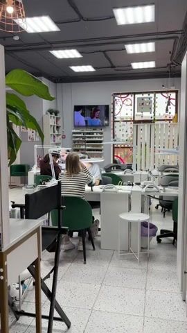 This media file captures the interior of a brightly lit nail salon in Imeni Morozova, Russia. The scene is dominated by white tables and chairs, with multiple nail technicians and clients engaged in manicures. A large flat-screen television mounted on the wall displays a scene from a television show or movie, featuring several actors. 

The salon is well-organized, with shelves lined with bottles of nail polish and other beauty products. Large potted plants add a touch of greenery to the space. The overall atmosphere appears relaxed and professional, with clients focused on their treatments. Natural light streams in from a window on the right, adorned with colorful circular decorations. The time of day is likely daytime, given the lighting. No visible text is present in the media file itself.