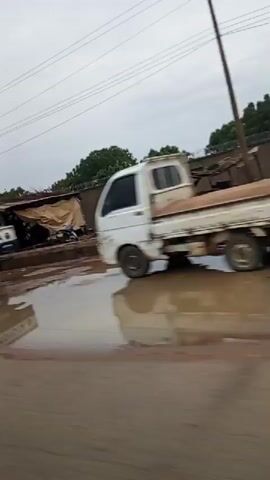 The video shows a blurry, shaky view from a moving vehicle. The scene outside appears to be a road with puddles and debris. Several vehicles are visible, including a white truck with a blue stripe, a red bus, a blue dump truck, a black car, and a dark blue hatchback. The environment seems to be an urban or semi-urban area with buildings and trees in the background. The sky is overcast.