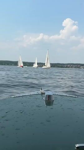 The video shows a first-person view from a boat moving across a body of water. The boat appears to be a motorboat, with a wooden deck and a metallic fixture at the front of the deck. Several sailboats are visible in the distance, with white sails against a blue sky and some clouds. The water is choppy, and there are small droplets of water on the surface of the boat's deck. The horizon is lined with trees and buildings, suggesting a shoreline. The scene appears to be a regatta or a sailing event. The camera is relatively steady, indicating smooth movement of the boat.