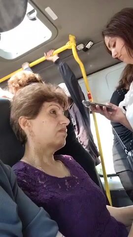 This is a close-up, first-person perspective video taken from inside a bus. In the foreground, a man wearing a gray cap and a dark shirt is visible. His face is turned downwards and slightly to the side. In the mid-ground, an older woman with short brown hair and a purple lacy top is seated and looking to her left. Behind her, a younger woman with long dark hair, wearing a white top, is holding a smartphone. The interior of the bus is visible, with yellow grab poles and seats. The lighting suggests it is daytime. The video captures a brief, candid moment of people on public transportation.