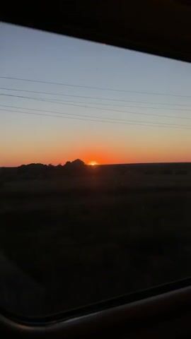 This video shows a sunset viewed from the window of a moving vehicle. The sun is low on the horizon, casting a warm orange glow across the sky. The landscape outside consists of fields and scattered trees, with utility poles lining the horizon. The interior of the vehicle is visible in the foreground, with the window frame and part of a seat. The scene is captured as the vehicle travels, indicated by the slight blur and changing perspective. The overall mood is tranquil and reflective, characteristic of a sunset journey.