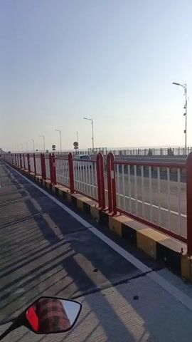 A first-person perspective video shows a motorcycle or scooter driving across a bridge. The sky is clear and blue, with the sun visible in the upper right portion of the frame. The bridge has a red and white railing along the side, and streetlights are spaced along the bridge. A few other motorcycles or scooters can be seen in the distance. The road surface appears to be asphalt, with white lines marking lanes. The sun casts long shadows on the road, indicating it is likely early morning or late afternoon.