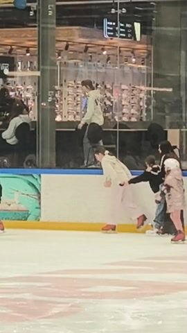 The video shows a group of people ice skating indoors. There are several people on the ice, some appear to be helping others, perhaps those who are new to skating. A young child in a pink coat and white helmet is also on the ice, being assisted by an adult. The environment appears to be an indoor skating rink, possibly within a shopping center or mall, as retail shops are visible in the background behind a glass barrier. There are colorful decorations along the edges of the rink. The overall scene suggests a recreational activity enjoyed by people of various ages.