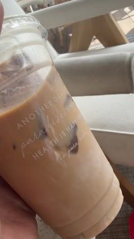 A hand holds a clear plastic cup filled with an iced coffee drink and ice cubes. The cup has white text printed on it that reads "ANOTHER DAY IN paradise WHERE HEALTH IS HAPPINESS xo pura vida." A straw is inserted into the cup. In the background, a white upholstered chair with wooden accents is visible. To the left of the chair is a small round table with a blue block displaying the number 27. Sunlight streams into the scene, suggesting an outdoor or well-lit indoor environment. The video shows the cup being held and then placed slightly to the side, revealing more of the background.