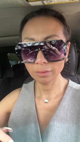 The video shows a close-up of a woman wearing large, angular sunglasses with a patterned frame. She is sitting in a car, with the interior visible behind her. She is wearing a grey vest over a white and black striped top. She has a thin silver necklace with a circular pendant. Her hair is pulled back. In the video, she moves her hand to her face, adjusting her sunglasses and then flashes a peace sign with her fingers. The reflection in her sunglasses shows another person, possibly the one filming, also wearing sunglasses.