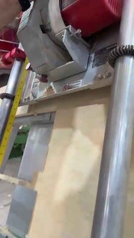 The video shows a man cutting a piece of plywood with a vertical panel saw. The saw has a red motor and a silver frame with two vertical chrome poles. The man is using his left hand to operate the saw and holds a black hand grip. The plywood is light-colored and positioned vertically. A yellow measuring tape is visible on the side of the saw. The environment appears to be a workshop or construction area. After the cut, the man lifts the black grip to adjust the saw.