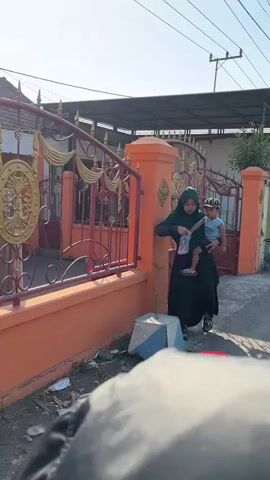 A woman wearing a green hijab and a long black dress walks by a fence carrying a baby. The baby is wearing a blue shirt and a cap. They are walking on a paved surface next to a residential building with an orange wall and a metal gate. The sky is clear and bright.