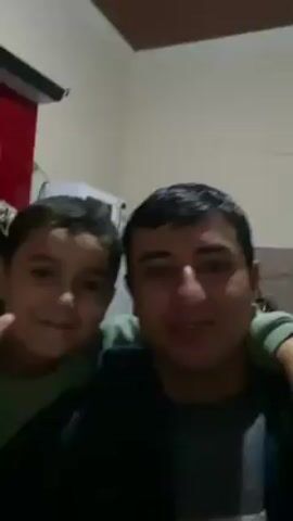 A man and a young boy are in a video. They are both smiling. The man is wearing a dark jacket and the boy is wearing a green shirt. The boy is holding up two fingers in a peace sign. They appear to be indoors, possibly in a bedroom. There is a red curtain and a white air conditioning unit in the background.