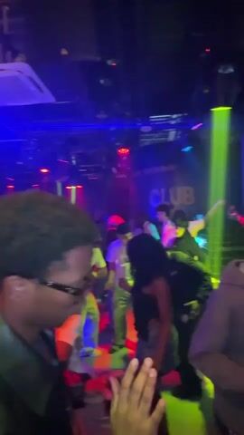 Inside a crowded, dark nightclub, a man wearing glasses and a dark jacket is seen from the side. The background reveals other individuals dancing under red, green, and purple stage lights. A sign reading "BAR - RESTAURANT CLUB 307" is visible in the background. As the camera shifts, a person in a green hat passes by. Throughout the video, people are seen dancing and socializing in the vibrant, bustling atmosphere of the club. The camera's movement is shaky.