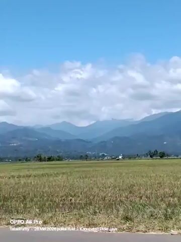 The video shows a rural landscape. There's a road in the foreground. Beyond the road is a large field with dry, yellow grass. In the distance are mountains under a blue sky with scattered white clouds. A motorcycle with two people, one wearing a white hijab, drives past the camera. The scene is bright and appears to be taken during the day. The bottom of the screen has text "OPPO A6 Pro", "fataelrumy 28 Mei 2026 pukul 11:41 Kabupaten Pidie Jaya".