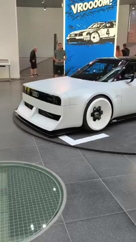 The video shows a white Audi concept car with distinctive white wheels and a black roof. The car is on a rotating display platform in a museum or showroom. The background features a large comic book-style mural with the word "VROOOOM!" and an image of a race car. Several people are visible in the background, including two men standing near the mural and another person on the right side of the frame looking at the car. The floor is tiled with a grey and white pattern, and a circular glass viewing area is visible in the foreground.