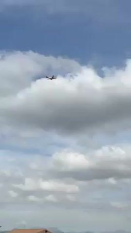 The video shows an airplane silhouetted against a cloudy sky. The plane appears to be at a high altitude and is flying away from the viewer. The sky is a mix of blue and white, with large, puffy clouds dominating the view. The ground is visible in the lower part of the frame, showing what seems to be a building or structure, with some greenery.