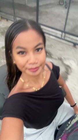 The video is a moving first-person perspective shot of a young woman with long dark hair wearing a black off-the-shoulder top and a gold necklace. She smiles at the camera as she moves through a green, possibly rural, environment with tall grass and banana trees visible in the background. The camera appears to be attached to the front of a moving vehicle, likely a scooter or motorcycle.