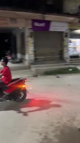 A nighttime street scene unfolds with a camera in motion, capturing a motorcycle in the foreground with its red taillight illuminated. Behind it, a person rides a bicycle, their attire including a green shirt with a pattern and checkered pants. The bicycle's front wheel is visible, and the rider appears to be looking towards the left. In the background, several shops line the street. One shop has a brightly lit interior displaying shelves of items. Another shop is partially visible with a yellow facade and signs. A few other individuals and motorcycles are present in the distance, contributing to the bustling yet somewhat dim atmosphere of the street. Debris, including plastic bags and paper, is scattered on the ground along the roadside.