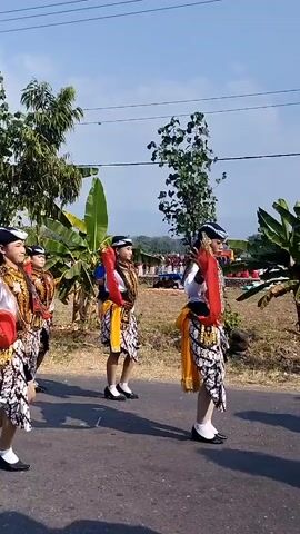 A group of young people, appearing to be predominantly female, are walking in a procession outdoors on a paved road. They are dressed in traditional clothing, characterized by white shirts, ornate gold-colored vests, patterned sarongs or skirts, and headwear. Some are also adorned with red sashes or streamers that they are holding or that trail from their garments. They are walking in a line, facing to the left of the frame, and appear to be moving forward. The environment includes trees and foliage on the left side of the road and a more open, possibly rural landscape with tents or temporary structures in the background on the right. The sky is visible and appears to be clear and blue. The lighting suggests it is daytime, possibly morning or afternoon.