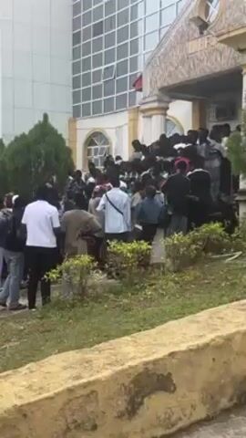 A large group of people is standing on and around the steps of a building. The building is light-colored and appears to be some kind of public or institutional building. There are windows on the upper floors. The area in front of the building is grassy with some small bushes. The people are dressed in a variety of casual clothing. Some are moving, while others are standing and talking. The overall impression is of a crowded scene, possibly an event or gathering.