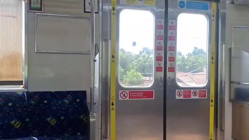 The camera looks at a closed train door as it travels. The door is stainless steel and has a window on each side, showing the landscape outside the train. The landscape includes residential homes, trees, and other greenery. The door has yellow vertical strips on each side and warning signs affixed to it. The interior of the train is visible, with a blue patterned seat on the left side and a blank sign space above it.