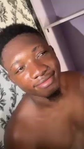 A shirtless young man with short black hair is recording a video. He has a tattoo on his left forearm. He looks up at the ceiling and then smiles at the camera. The room has a lavender ceiling and a white shelf. The wall behind him is covered in a white and gray patterned wallpaper. The man is wearing green shorts.