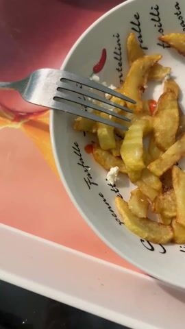 The video shows a plate of fries topped with small pieces of white cheese. A fork rests on top of the fries. The plate has Italian pasta names printed around the rim, such as "tortellini" and "penne," along with a red chili pepper illustration. The plate sits on a white tray that has a peach-colored surface with a yellow floral design. The camera briefly loses focus and then returns to the plate. The speaker says they have almost finished their dinner and tea and it is time for "the peel," presumably referring to the food preparation or disposal of the leftovers.