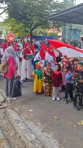 A group of people, including children and adults, are participating in a parade or procession. Many of the participants are wearing traditional Indonesian clothing and some are holding Indonesian flags. The children are dressed in various colorful outfits, and some appear to be dressed as soldiers or scouts. Adults are also present, some of whom are wearing hijabs and holding drums. The procession is taking place outdoors on a paved street, with trees and buildings visible in the background. The overall atmosphere appears festive, suggesting a celebration or a cultural event.