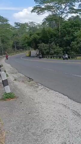 A yellow truck drives on a winding road surrounded by trees and vegetation. A scooter follows closely behind the truck. The road has a yellow line down the center and a gray shoulder. There are also a few other scooters visible on the road. The sky is cloudy.