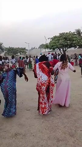 The video shows a group of people dancing outdoors in a circle. The camera is held at ground level and follows the movement of the dancers. Most people are wearing brightly colored traditional clothing. Some of the dancers are holding hands. The ground is sandy, and there are trees, buildings, and a lamppost visible in the background. The sky is overcast.