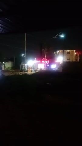 This video captures a nighttime scene of a small, brightly lit structure with red and green neon signs. The structure appears to be a roadside establishment or kiosk, possibly a food vendor or small shop. The lighting is intense, creating a high-contrast image with deep shadows. In the background, there are glimpses of buildings and streetlights, suggesting an urban or semi-urban environment. The foreground is dark and appears to be unpaved, possibly a dirt or grassy area. There are no people visible in the video. The overall impression is one of a vibrant, commercial spot operating late at night.
