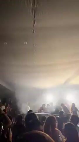 The video shows a live music event inside a rectangular room with a low, flat ceiling. The focus is on a DJ setup, likely comprising turntables and electronic equipment, positioned on a table at the far end of the room. The DJ and their associates stand behind the setup. In front of them is a crowd of people, most of whom are visible only from the shoulders up. Some audience members are raising their arms in the air, clearly enjoying the music. The lighting is dynamic, shifting between bright flashes and periods of darkness, creating a strong contrast and accentuating the party atmosphere. Additional stage lighting is visible behind the DJ, contributing to the overall spectacle. There is also visible smoke or fog in the air, which contributes to the ambiance.