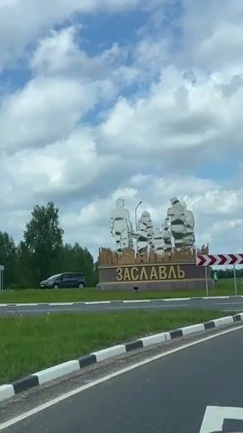 The video shows a drive-by view of a sign marking the entrance to Zaslavl, Belarus. The sign features chrome figures of a family standing on a brown base with the city's name in yellow Cyrillic lettering. The sky is blue with scattered clouds, and the road is bordered by green grass and trees. There is also a red and white directional sign next to the city name. A black car is visible parked in the background.