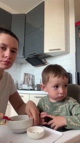 A woman is seen spoon-feeding a young child in a bright, modern kitchen during the daytime. The woman, with dark hair tied back in a bun and wearing a white t-shirt, sits on the left, holding a white bowl filled with a light-colored, lumpy food (possibly scrambled eggs or porridge) and a spoon with a red handle. Her expression is focused as she tends to the child.

The child, a toddler with short brown hair, is seated in a high chair with a white tray, wearing a green long-sleeved shirt with light-colored dinosaur patterns. There are visible smudges of food, possibly chocolate or a dark sauce, around the child's mouth and on their face, indicating a messy but engaged meal. The child looks intently towards something off-camera to the right, with wide, curious eyes. A smaller, empty light brown bowl rests on the child's tray.

The background reveals a clean, contemporary kitchen with a white subway tile backsplash. The upper cabinets are a combination of dark gray and off-white, featuring sleek horizontal handles. A sink area with a faucet and some kitchen items is visible between the cabinets, and a dark appliance, possibly an oven hood or microwave, is mounted above the sink.