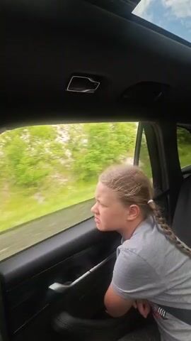 The video, shot from inside a moving car, shows a young girl looking out the passenger side window. She has light-colored hair styled in two braids. The car appears to be driving on a road with green vegetation visible outside the window. Through the window, other vehicles can be seen on the road. The interior of the car is mostly dark, with a sunroof visible at the top of the frame. The girl seems contemplative as she watches the passing scenery. Music is playing in the background.