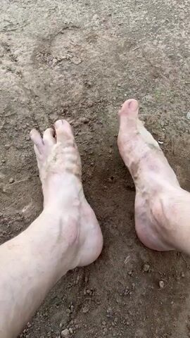 The video shows two bare feet resting on the ground. The ground is dirt, and there is small debris scattered around. The feet are light-skinned and have visible veins. There is dirt on the feet. The toes are visible, and the nails are not painted. There are no other objects or people visible in the video.