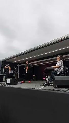 A live band of three people performs on an outdoor stage under a cloudy sky. A woman on the left sings into a microphone, wearing a black outfit. In the middle, a man plays the trumpet, dressed in black with a hat and red shoes. On the right, a man sits at a keyboard, wearing a patterned shirt and jeans, playing along. The stage is inside a trailer, with speakers and equipment visible.