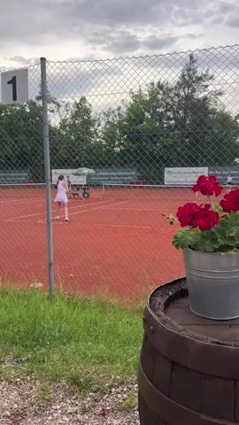 A person wearing a white tennis outfit is playing tennis on a clay court. They are on the left side of the court, facing the net. The person is in the motion of hitting a tennis ball. There is a tennis net in the middle of the court, and another person is visible on the other side of the net, also playing tennis. A fence surrounds the court, and a sign with the number "1" is attached to a fence post. In the foreground, there is a barrel with a metal bucket of red flowers on top. The sky is cloudy, and trees are visible in the background.