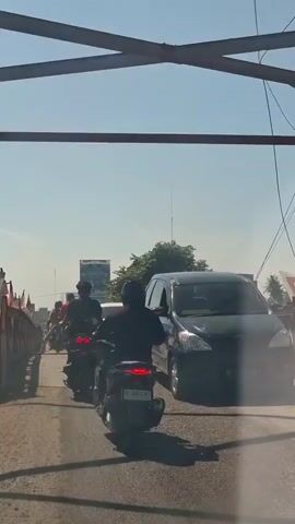 The video shows a view from the perspective of someone on a motorcycle, looking forward and slightly upward. The camera is angled down, and a portion of the motorcycle handlebar and front wheel is visible. The scene depicts a traffic jam on a bridge. Several motorcycles and cars are visible, moving slowly. The motorcycles in front are also carrying passengers. In the background, there are trees, buildings, and flags. The sky is clear and blue, indicating a sunny day. The bridge appears to be made of metal and has railings. The road surface is asphalt. The overall atmosphere is one of a busy, everyday commute.