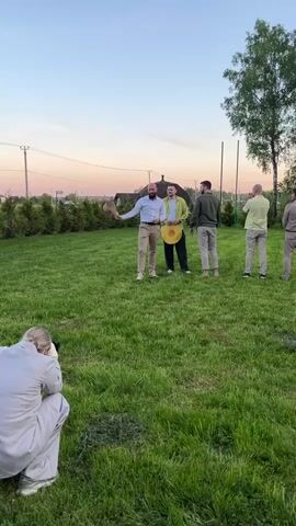 The video shows a photographer crouching in the foreground, taking pictures of four men standing on a grassy lawn. The men are dressed in light-colored, casual clothing, with three of them holding hats. One man throws his hat in the air, adjusting his shirt collar as it falls. Behind the men, there's a small house, a line of bushes, and power lines under a colorful sky. The environment appears to be a yard or garden.