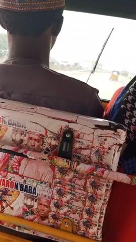 A person is riding in the back of a yellow auto rickshaw (tuk-tuk).  The person is wearing a brown shirt and a decorative hat. The back of the driver's seat is covered with a white patterned covering with images of a man and the text "Yaron Baba". The rickshaw is moving down a road. The driver is on a cell phone. Other vehicles can be seen outside the rickshaw. There is another passenger to the right.