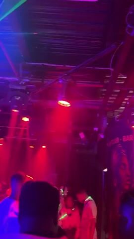 A vertical video taken from a low angle captures a busy nightclub scene. The space is dimly lit, with blue and red lights illuminating the area and creating a lively atmosphere. Numerous individuals can be seen in the club, with some moving to the music and others conversing. The camera pans slowly to the right, showing more of the club's interior and the diverse crowd enjoying themselves. On the far wall, a large banner with "CLUB 007" written in bold letters is visible, adding to the vibrant decor. The video concludes with the camera focused on the club's interior, providing a final glimpse of the active and engaging environment.