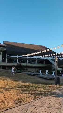 The video pans from left to right, showcasing an outdoor event with a clear blue sky. In the foreground, a cobblestone path leads towards a large building with a dark, sloped roof. The building is adorned with strings of white, round lanterns, creating a festive atmosphere. Beyond the building, a large gathering of people is seated at white-covered tables, suggesting a formal event or reception. The scene is further decorated with white umbrellas and tables set up outside the main building. A bicycle is parked on the cobblestone path. To the right, a body of water with reflections of the trees and sky is visible, bordered by a metal railing. The overall impression is of a beautifully set-up outdoor event on a pleasant day.