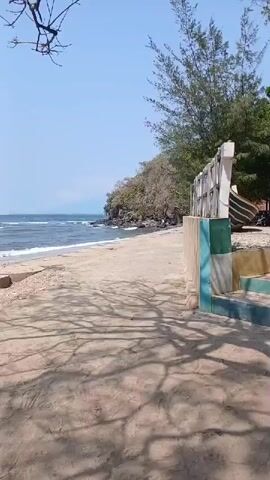 The video opens with a view of a beach with a calm sea and clear blue sky. The camera pans to the right, revealing a sandy path with shadows of trees cast upon it. To the right of the path, there is a concrete structure with colorful tiles and a white sign that is partially visible. Further along the path, a large tree with sparse branches can be seen. The beach itself has sand and some scattered rocks. In the distance, a rocky coastline with vegetation is visible. The sea has gentle waves lapping at the shore. The video continues to pan right, showing more of the sandy beach and the sea. The concrete structure continues, featuring circular concrete blocks and some rubble. The camera then moves slightly to the left, offering a wider view of the beach and the sea. The video ends with a close-up of the circular concrete blocks and rubble, with the sea in the background.
