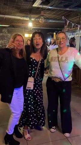 Three women are standing and dancing in a dimly lit indoor space with a tiled floor. The woman on the left is wearing a black blazer over a black shirt and white pants, with black ankle boots. The woman in the middle is wearing a black dress with white polka dots and a white crossbody bag. The woman on the right is wearing a white long-sleeved shirt with sparkly text on it and dark pants with white sneakers. They are all smiling and appear to be enjoying themselves. Green and other colored lights are projected onto them and the surroundings, suggesting a party atmosphere. In the background, there are some tables, chairs, and decorative elements, including what appears to be a menu board on the left wall. The ceiling has exposed beams and industrial-style lighting fixtures.