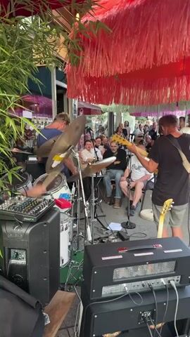 The video shows a band performing live outdoors. A drummer is visible on the left side of the frame, playing a drum kit. In the center, a guitarist is playing a yellow electric guitar and facing the audience. The audience is seated and standing, watching the band. The stage is set up outdoors, possibly on a patio or sidewalk, with bamboo plants on the left and red, fringed decorations hanging overhead. There are musical instruments and audio equipment, including amplifiers and a mixing board, visible on the stage. The overall atmosphere appears to be casual and lively.