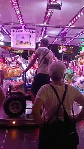 The video shows a carousel ride at what appears to be a fair or amusement park. The carousel has various themed carriages, including a pumpkin carriage, a tractor, and animal figures. People are seen standing on and around the carousel. In the foreground, a woman with gray hair and a black backpack is facing away from the camera, watching the ride. Lights and decorations are visible on the carousel's ceiling. The ride is in motion, with carriages spinning around a central axis. Some of the carriages appear to be designed for children, with animal seats and colorful designs. A screen displaying the words "Video Baby" is visible on the carousel's structure. The overall impression is of a lively and festive atmosphere typical of an amusement park.