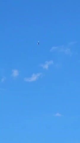 A tiny jet plane is captured in flight against a vast, clear blue sky with a few wispy white clouds. The camera appears to be stationary, looking upwards at the plane. The jet is the sole focal point of the video.