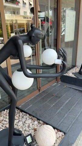 The video shows a close-up of black stylized sculptures on a sidewalk outside a building with glass doors. The sculpture in the foreground features a black arm holding a white sphere and a white card. To the right, a larger black sculpture of a human-like figure with extremely long limbs stands with two white spheres in its hands. The figure is positioned in a black planter filled with white pebbles. The building's glass doors reflect the surroundings. The ground is paved with dark rectangular tiles and gravel.