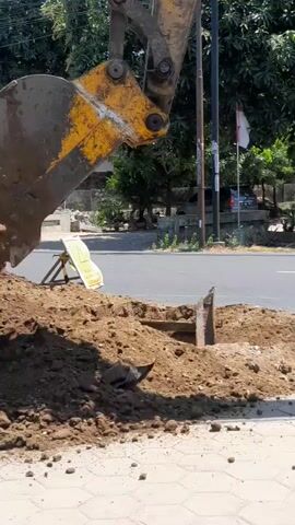 An excavator is digging a trench on a paved surface next to a road. The excavator's bucket is lowered into the dirt, and a pile of excavated soil is visible. A yellow and black sign with a warning symbol is placed near the trench. Cars and motorcycles are visible on the road in the background, and trees and buildings line the street. The scene appears to be an outdoor construction or repair site.